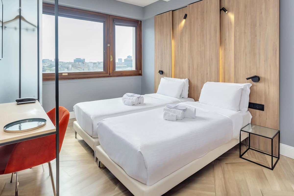 Ett hotellrum med två sängar, ett skrivbord, en stol och utsikt över staden genom stora fönster.