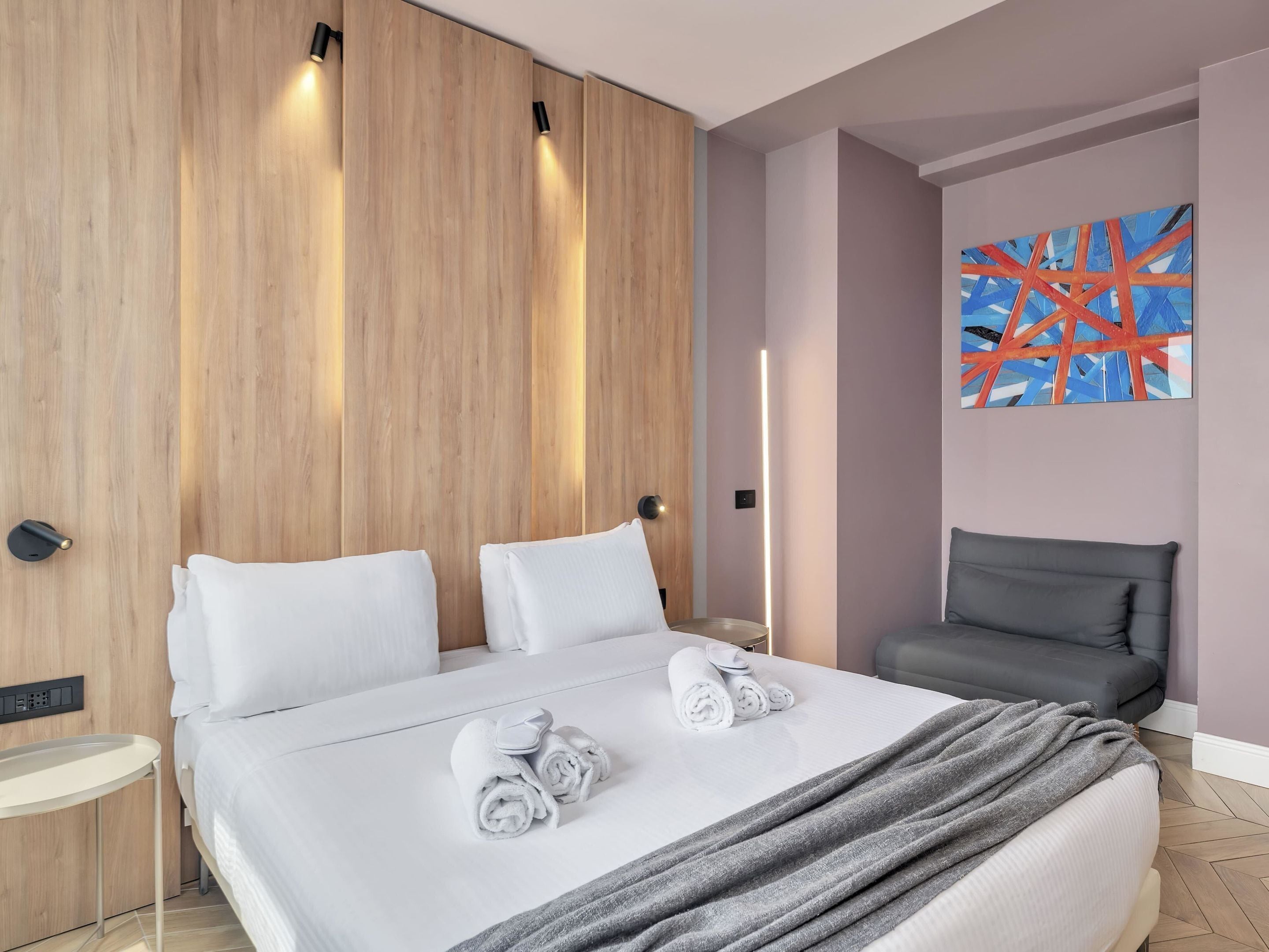 Ein modernes Hotelzimmer mit einem großen Bett, einem grauen Sessel, einem Gemälde an der Wand und einem Nachttisch mit einer Lampe.