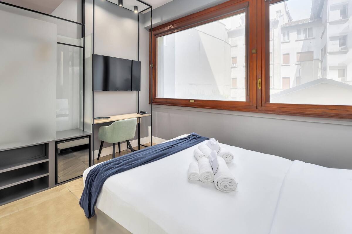 Ett hotellrum med en säng, ett skrivbord, en stol, en tv och ett fönster med utsikt över byggnader.
