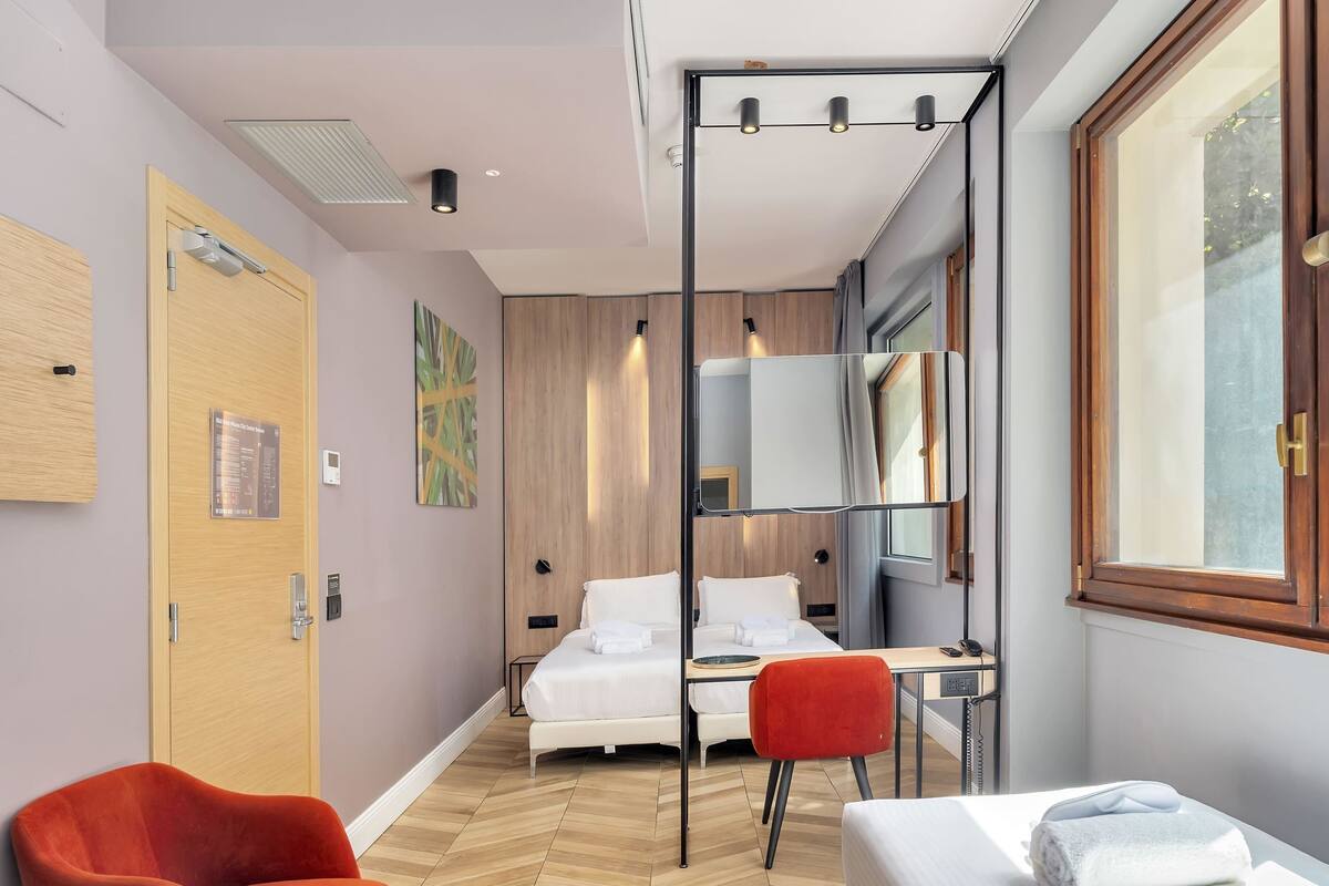 Ett modernt hotellrum med trägolv, en säng med vita sängkläder, en röd stol, ett skrivbord med en stol och ett stort fönster.