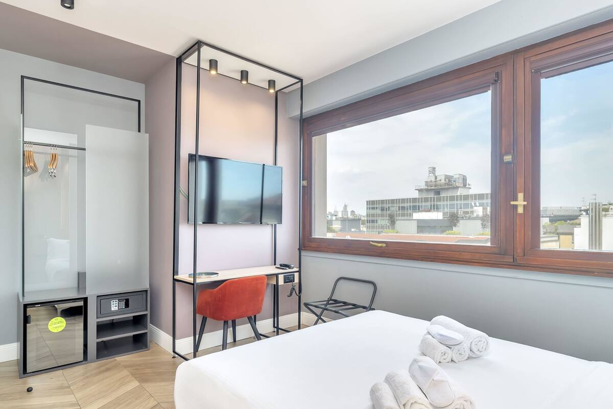 Ett hotellrum med en säng, ett skrivbord, en TV, en garderob och ett fönster med utsikt över staden.