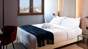Habitación de hotel con una cama grande, una silla roja, una mesita y vista a la ciudad por una ventana.