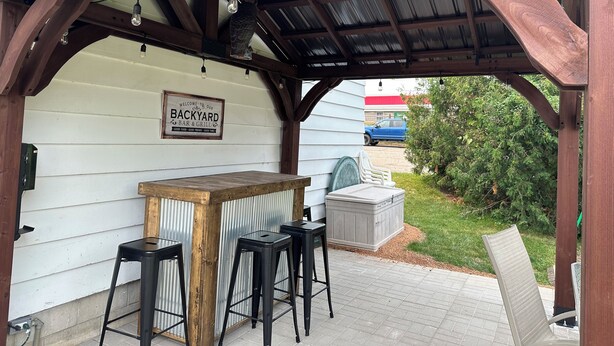 Un área de bar al aire libre con techo, que cuenta con una barra y taburetes de madera, un cartel que dice "BACKYARD BAR & GRILL", y vistas de un paisaje verde.