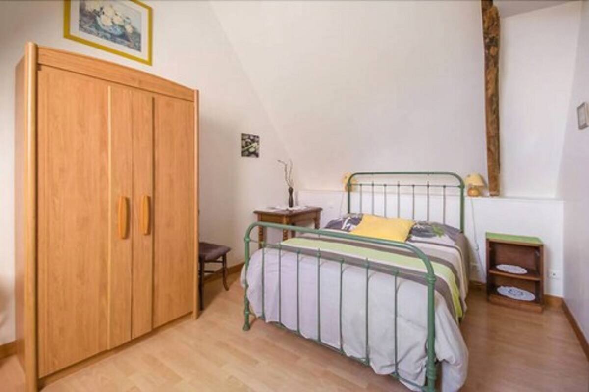 A bedroom with a wooden wardrobe, a single bed with a green metal frame, a bedside table, and a small shelf.