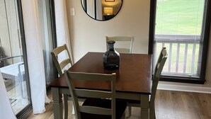 En matplats med ett träbord, fyra stolar, en hängande lampa, en spegel och utsikt över en grön gräsmatta genom ett fönster med persienner.