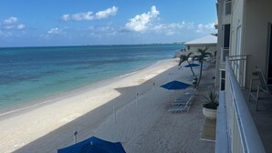 Une plage de sable blanc bordée d’une eau d’un bleu cristallin. Des chaises longues et des parasols sont disposés le long du rivage. Un bâtiment doté d’un balcon surplombe la plage.