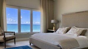 Une chambre avec un grand lit, une chaise, une table de chevet avec une lampe, et une vue sur l’océan grâce à de grandes fenêtres.
