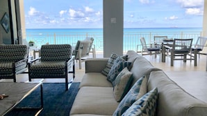 Un vaste espace de vie comprenant un grand canapé, des fauteuils à motifs et une table à manger. Un balcon doté d’une balustrade offre une vue sur une mer d’un bleu profond.