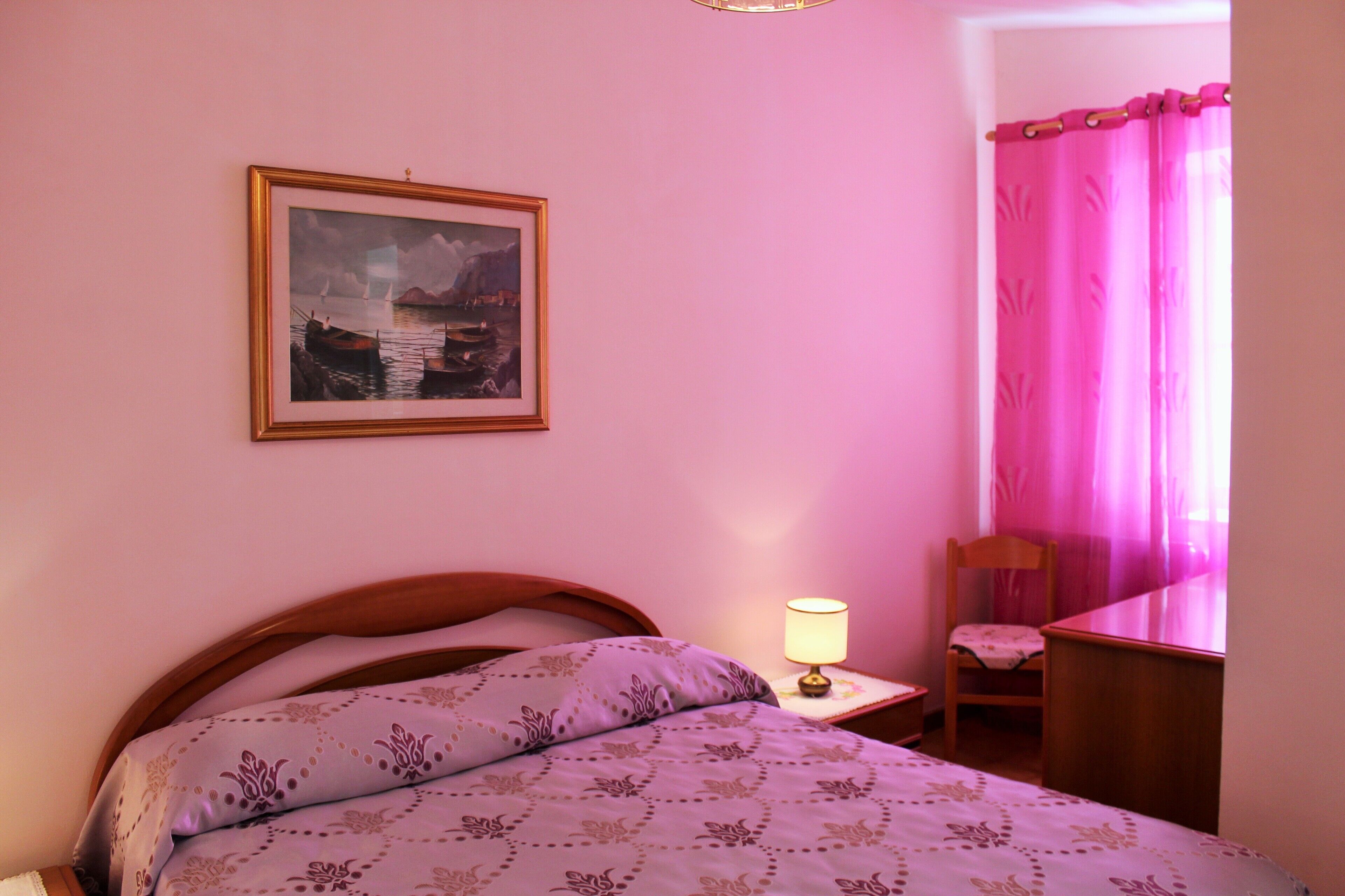 A bedroom with a bed, a chair, a nightstand with a lamp, a framed picture on the wall, and a pink curtain.