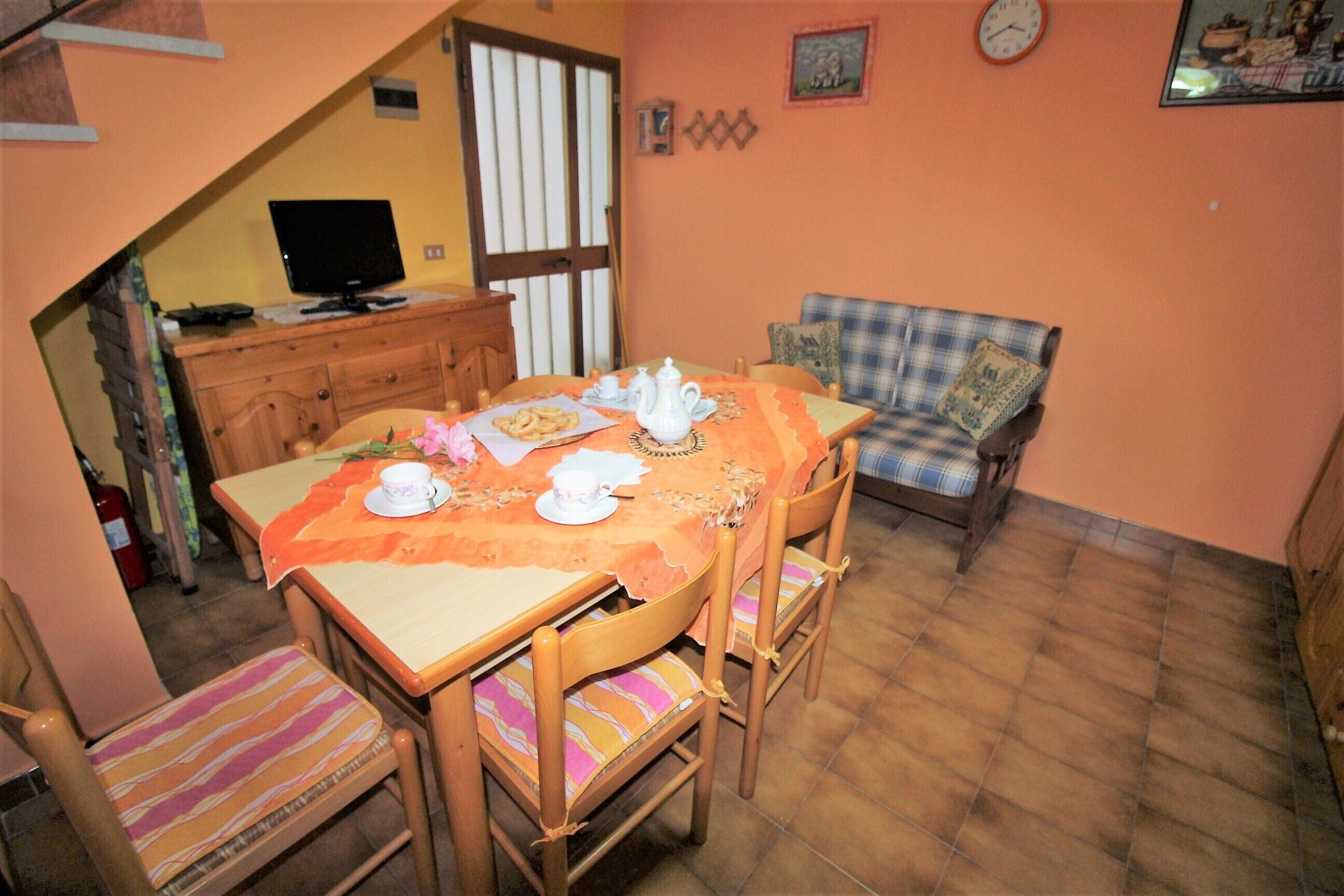A dining area with a table set for four, a television on a stand, and a sofa in the background.