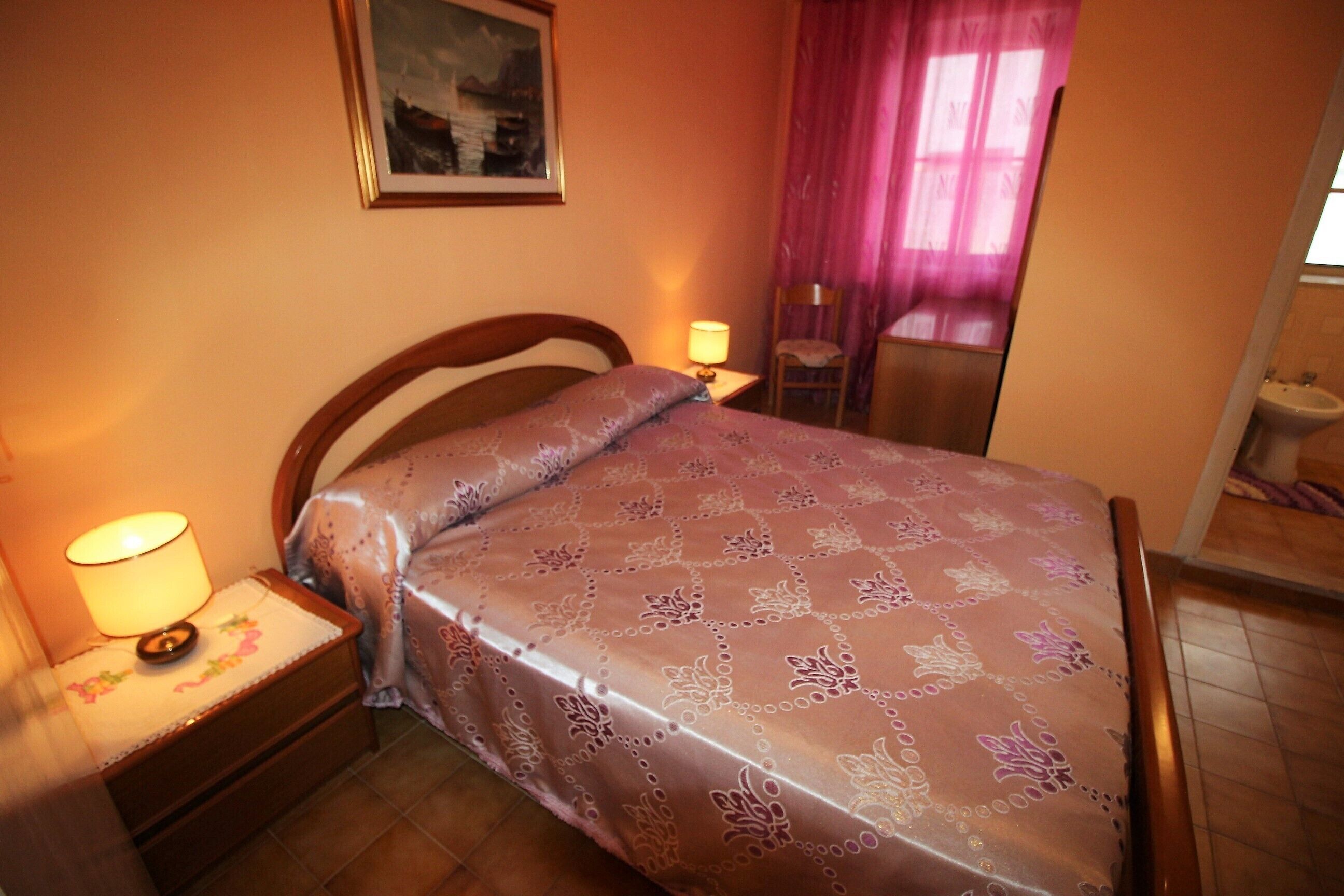 Quarto com cama, mesa de cabeceira, abajur, cadeira e um quadro na parede.