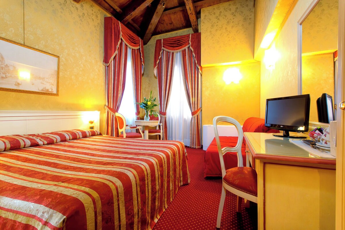 A hotel room with a large bed, a desk with a computer, a chair, and a small table with a vase of flowers.