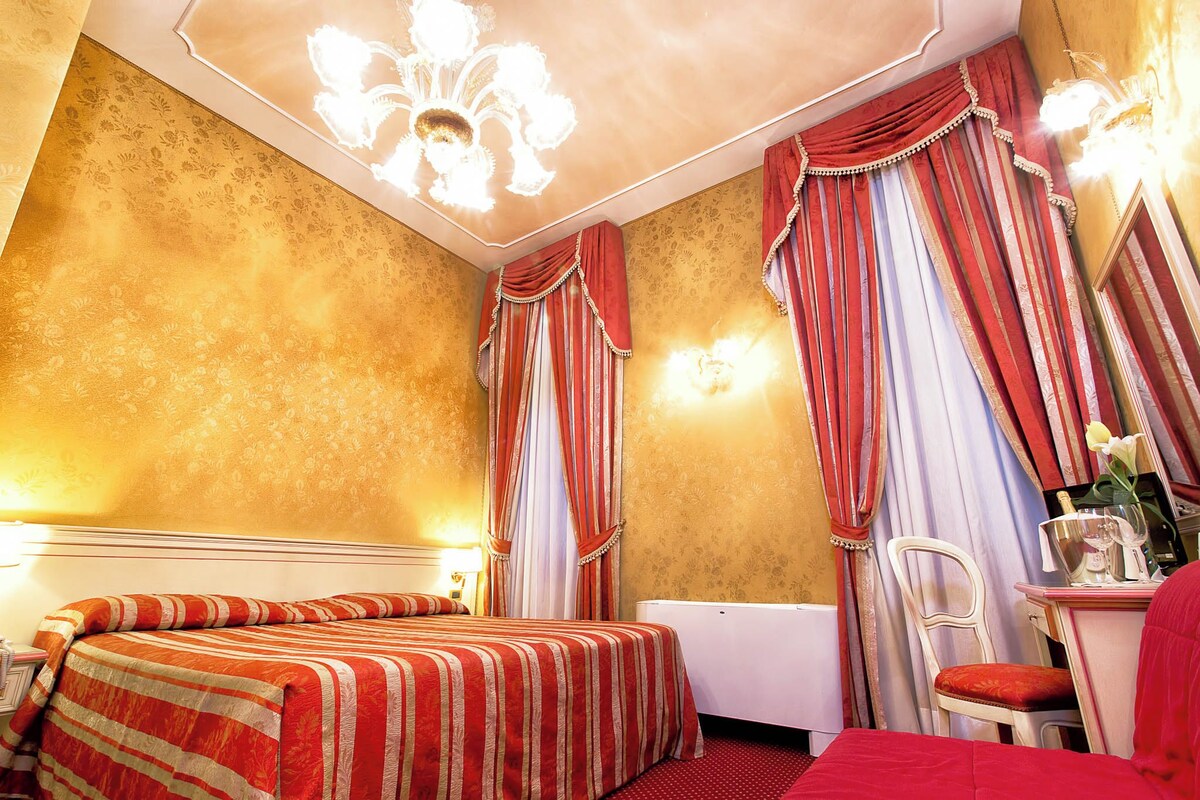 Habitación de hotel con cama, escritorio y silla. La habitación tiene una lámpara de araña, cortinas y un radiador.