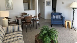 Ein Wohnzimmer mit Esstisch für vier Personen, einem blauen Sessel, einem gestreiften Sofa und einer großen Farnpflanze.