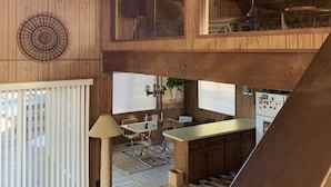A spacious loft-style room with a high ceiling, wooden beams, a dining area, and a kitchen with a refrigerator. - 1970s Architecture & Design Cabin - The Lone Pine (Frazier Park)