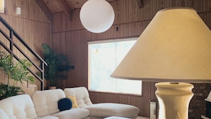 A cozy living room with a beige sofa, a coffee table with books, a white lamp, and wooden walls. - 1970s Architecture & Design Cabin - The Lone Pine (Frazier Park)