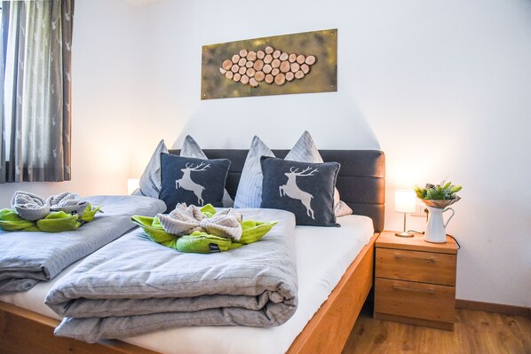 A neatly made bed with grey linens, a decorative pillow with a deer design, and a bedside table with a lamp and a plant.