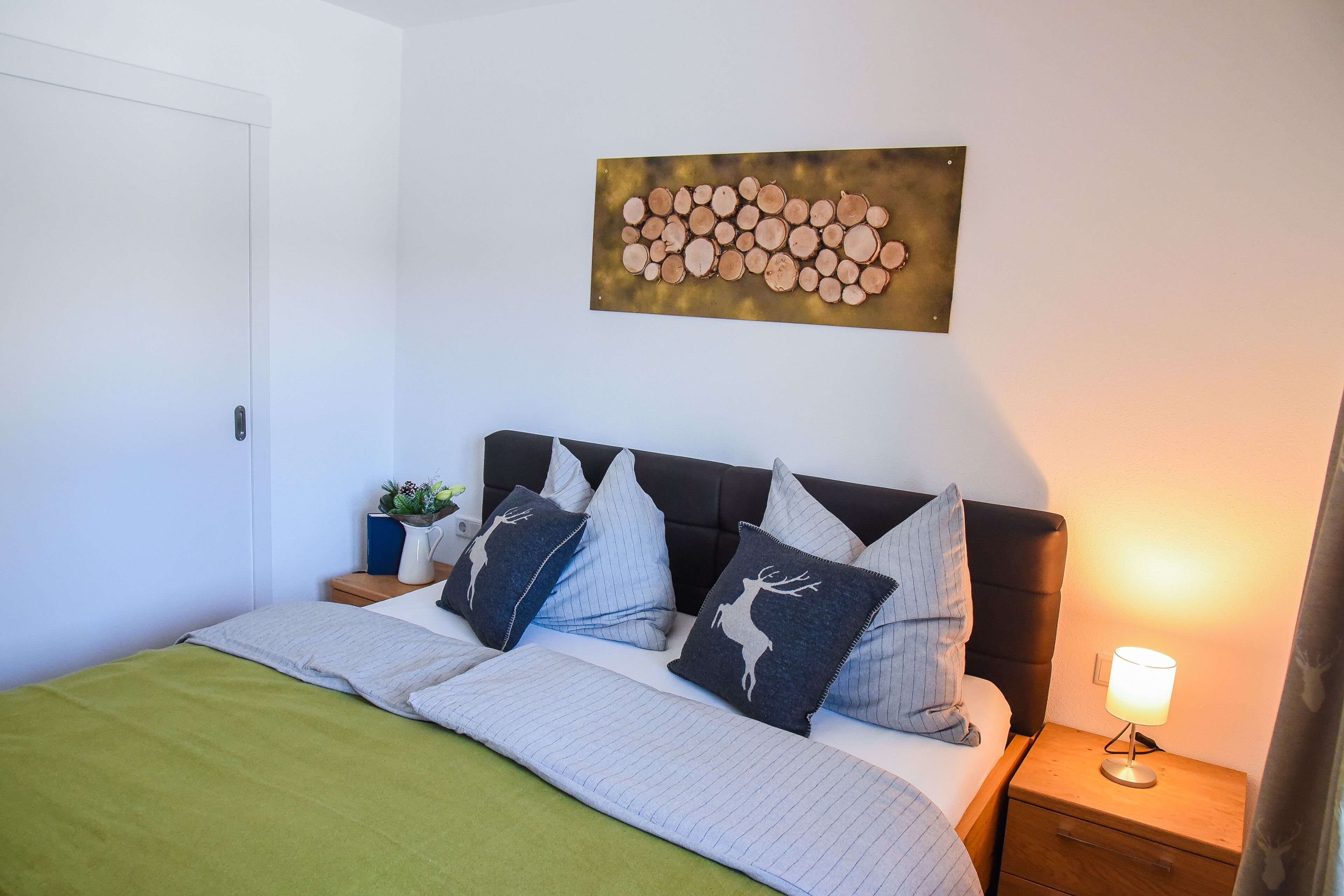 Una cama bien hecha con una manta verde, cabecera con varios almohadones, mesita de noche con lámpara y una obra de arte colgada en la pared.