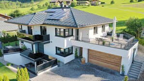 A modern two-story house with a flat roof, large windows, and a balcony with outdoor furniture.