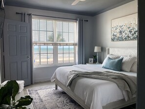A bedroom with a bed, a window with a view of the beach, a bedside table with a lamp, and a painting on the wall.