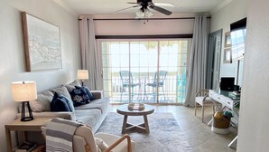 A living room with a sofa, armchair, coffee table, and a TV on a stand. There is a sliding glass door leading to an outdoor area with chairs. - Spectacular sunrise and ocean views! Award winning "Best Condo Rentals!" (Galveston)