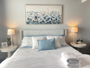 Een net opgemaakt bed met wit- en blauwe beddengoed, twee nachtkastjes met lampen, een lijst met bloemistkunst boven het bed, en een kleine potplant op een tafeltje.