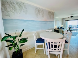 Een eetgedeelte met een ronde tafel en witte stoelen, een kamerplant en een muurschildering van een strand.