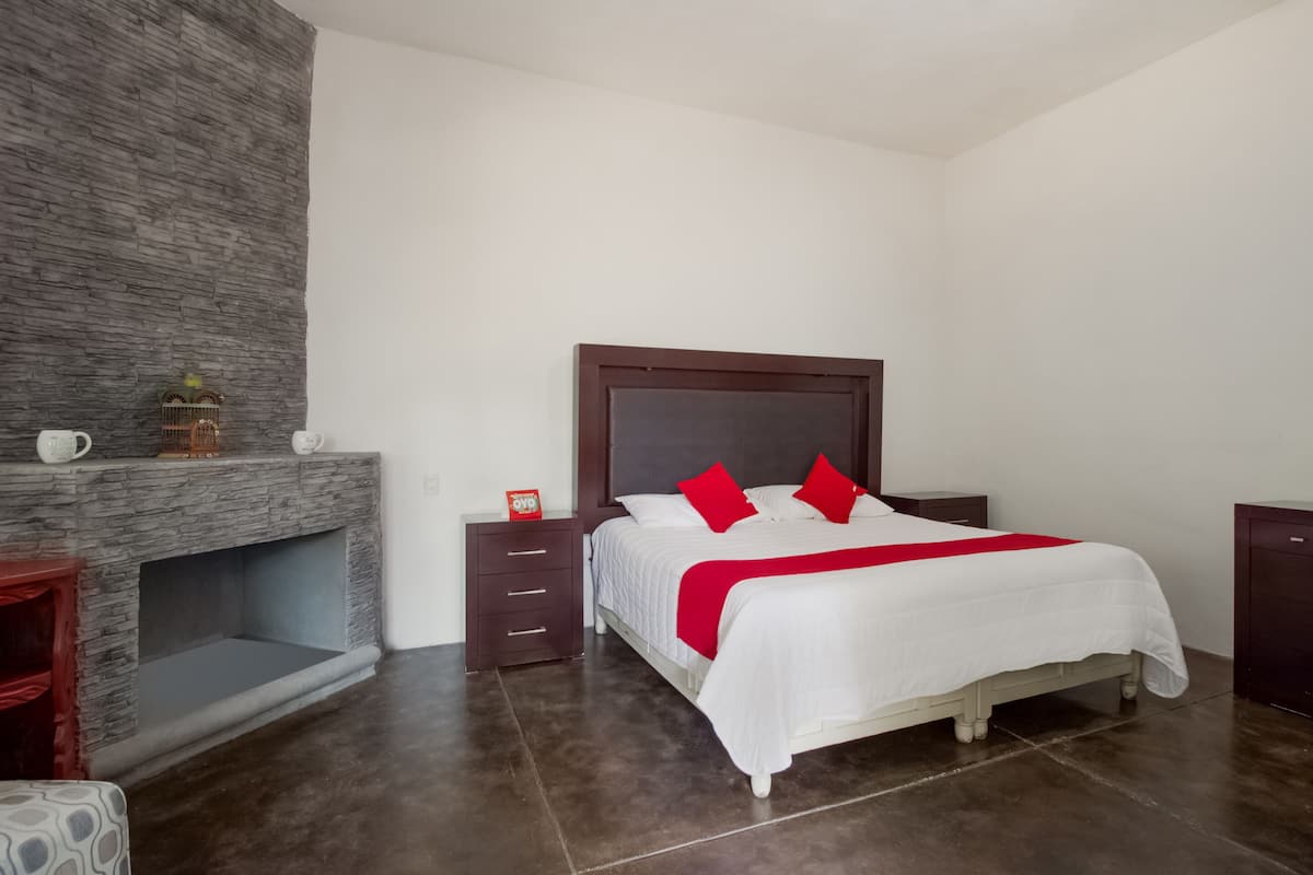 Un dormitorio con chimenea de piedra, una cama con ropa de cama blanca y almohadas rojas, una mesita de noche y un aparador.