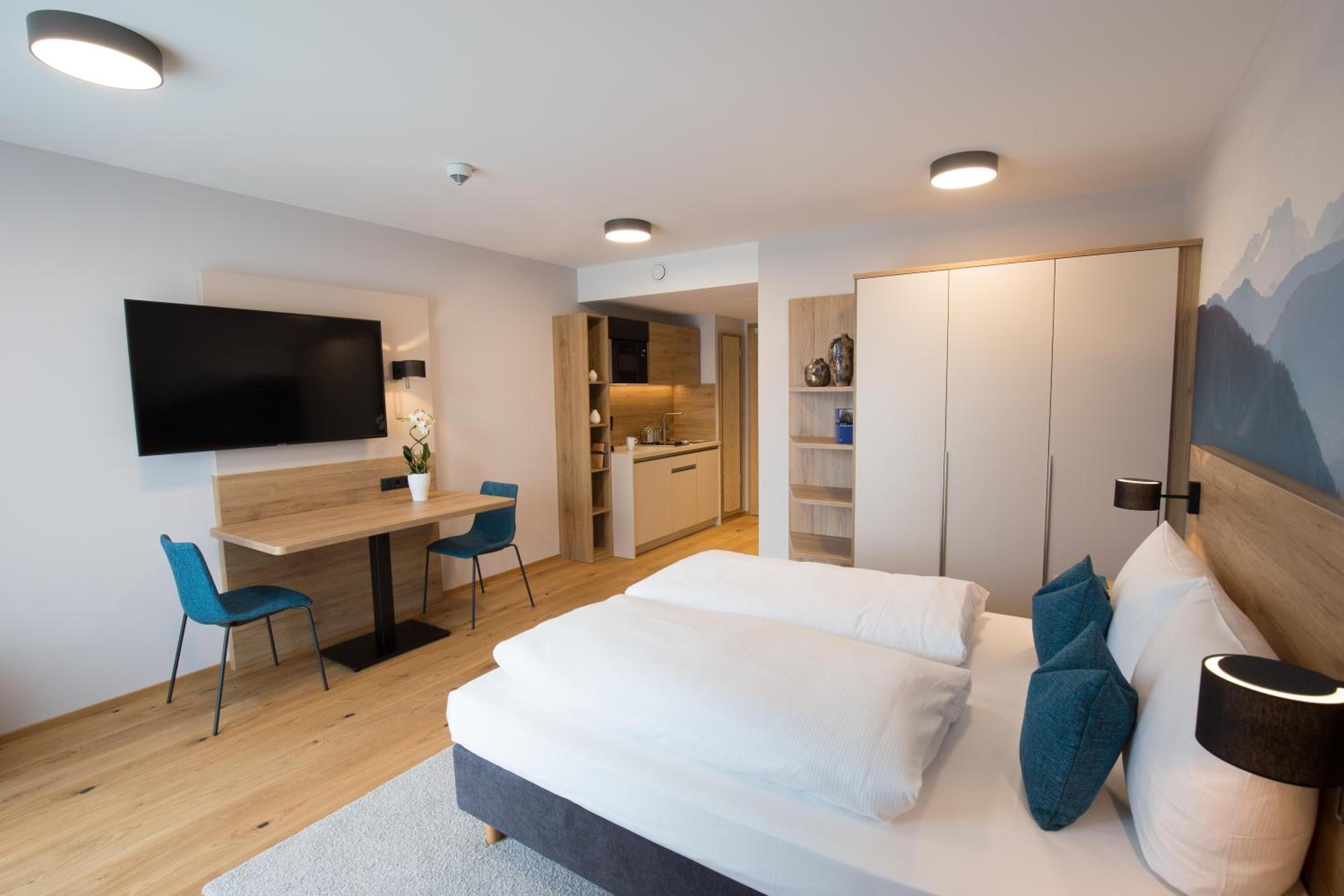 Una habitación de hotel moderna con cama, escritorio, silla, televisión y una zona de cocina.