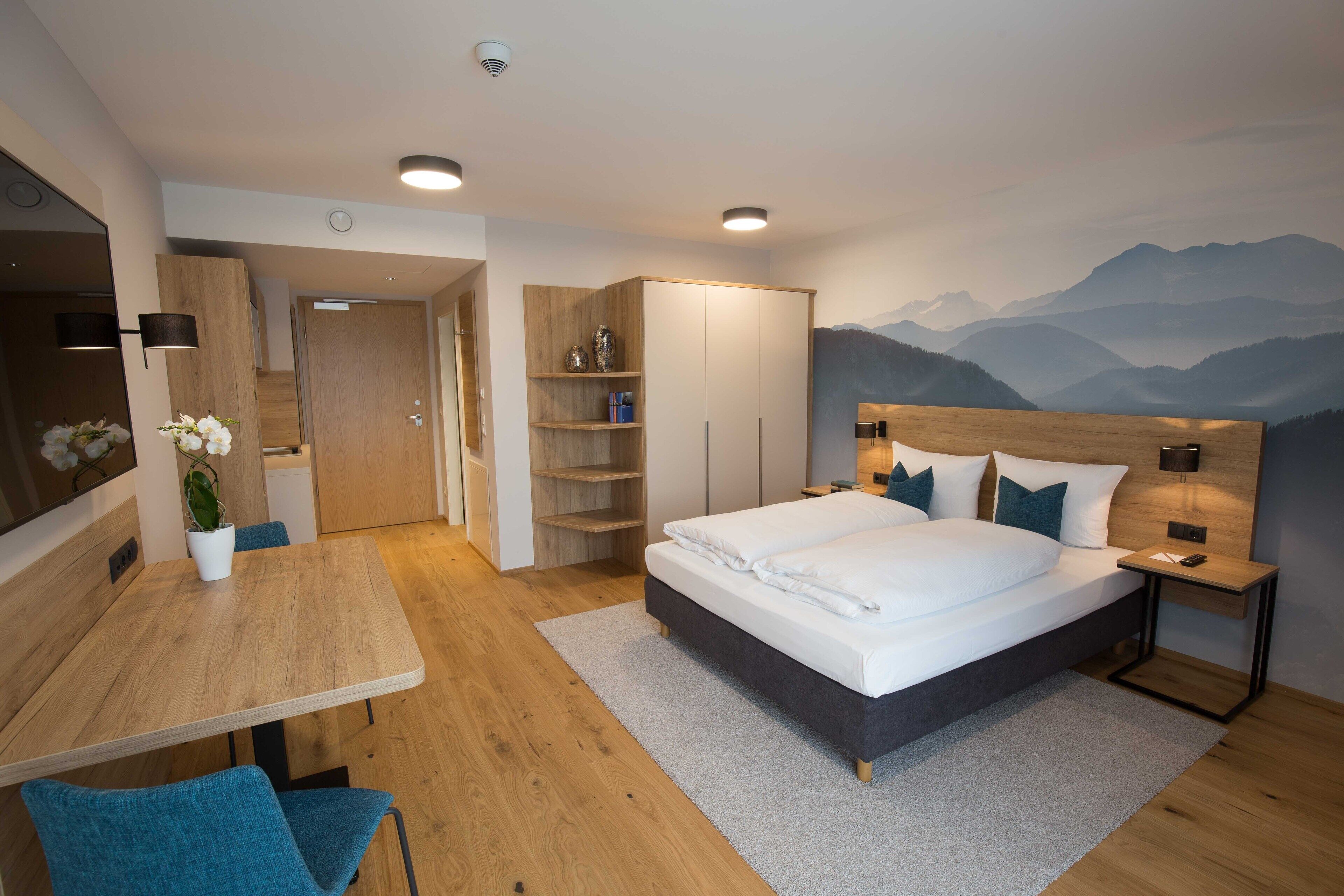 A modern hotel room with a large bed, a desk, and a chair. There is a wardrobe, a shelf with decorative items, and a wall-mounted lamp.