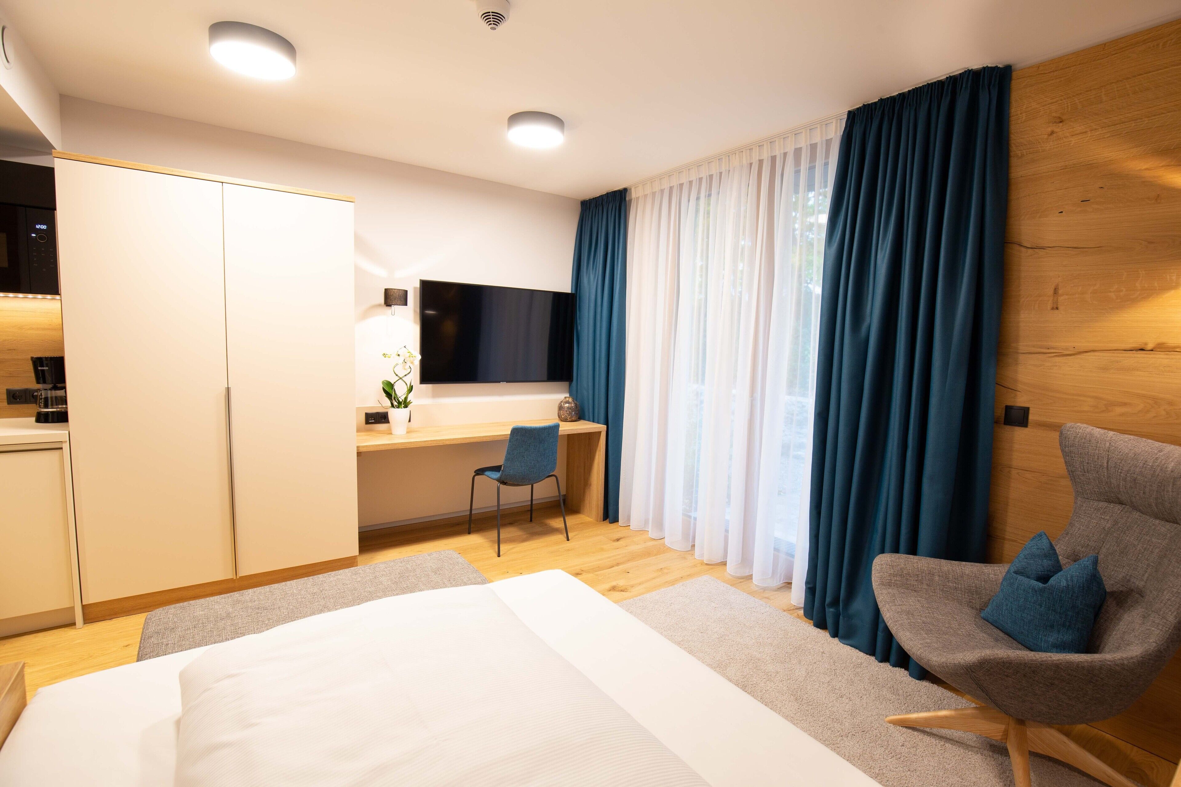 A modern hotel room with a bed, a desk with a chair, a TV, and a large window with curtains.
