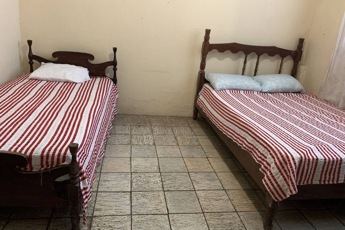 Two single beds with wooden frames and red and white striped bedding, in a room with tiled flooring and plain walls.