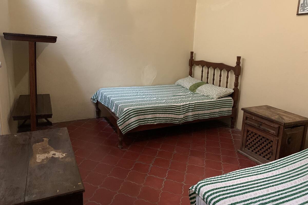 A small, dimly lit room with a single bed, a wooden nightstand, and a small table.