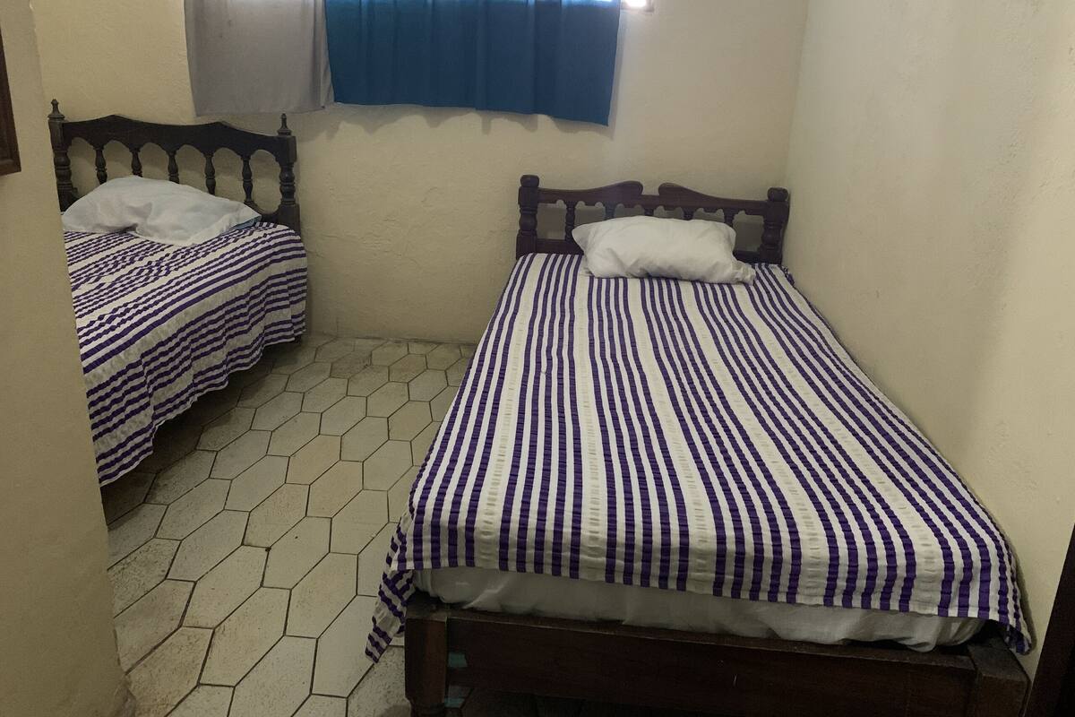 A small, clean room with two single beds, each with a wooden headboard and striped bedding. The room has a tiled floor and a window with blue curtains.