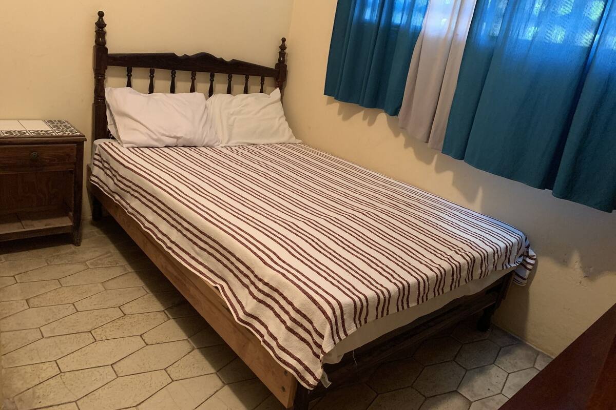 A single bed with a striped bedspread, a wooden nightstand, and a window with blue curtains.