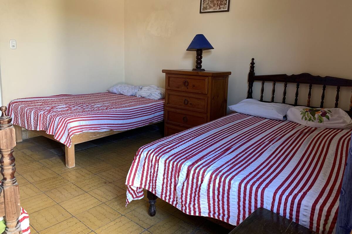 A room with two beds, each with a striped bedspread, a wooden dresser, and a lamp.