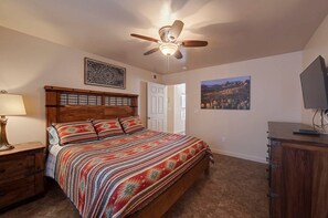 Une chambre à coucher avec un lit en bois, un ventilateur de plafond, un bureau avec un ordinateur et une porte donnant sur une autre pièce.