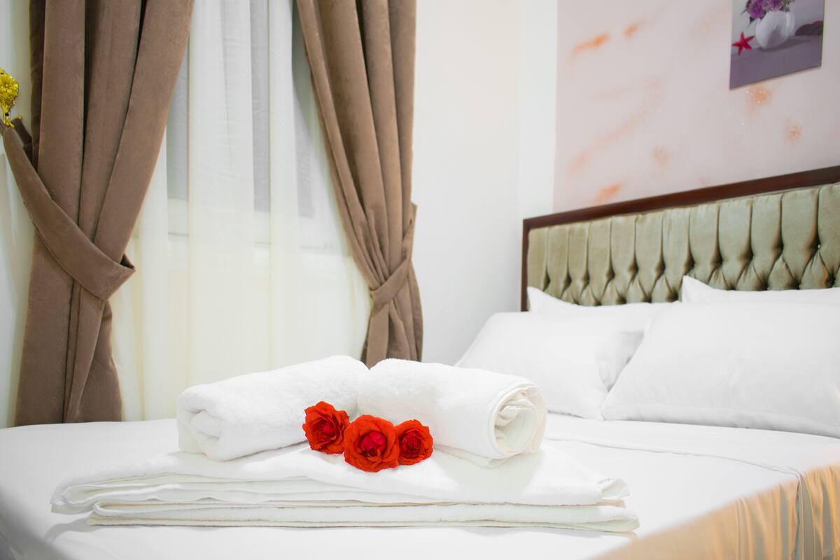 A neatly made bed with white linens, a rolled white towel, and three red roses.