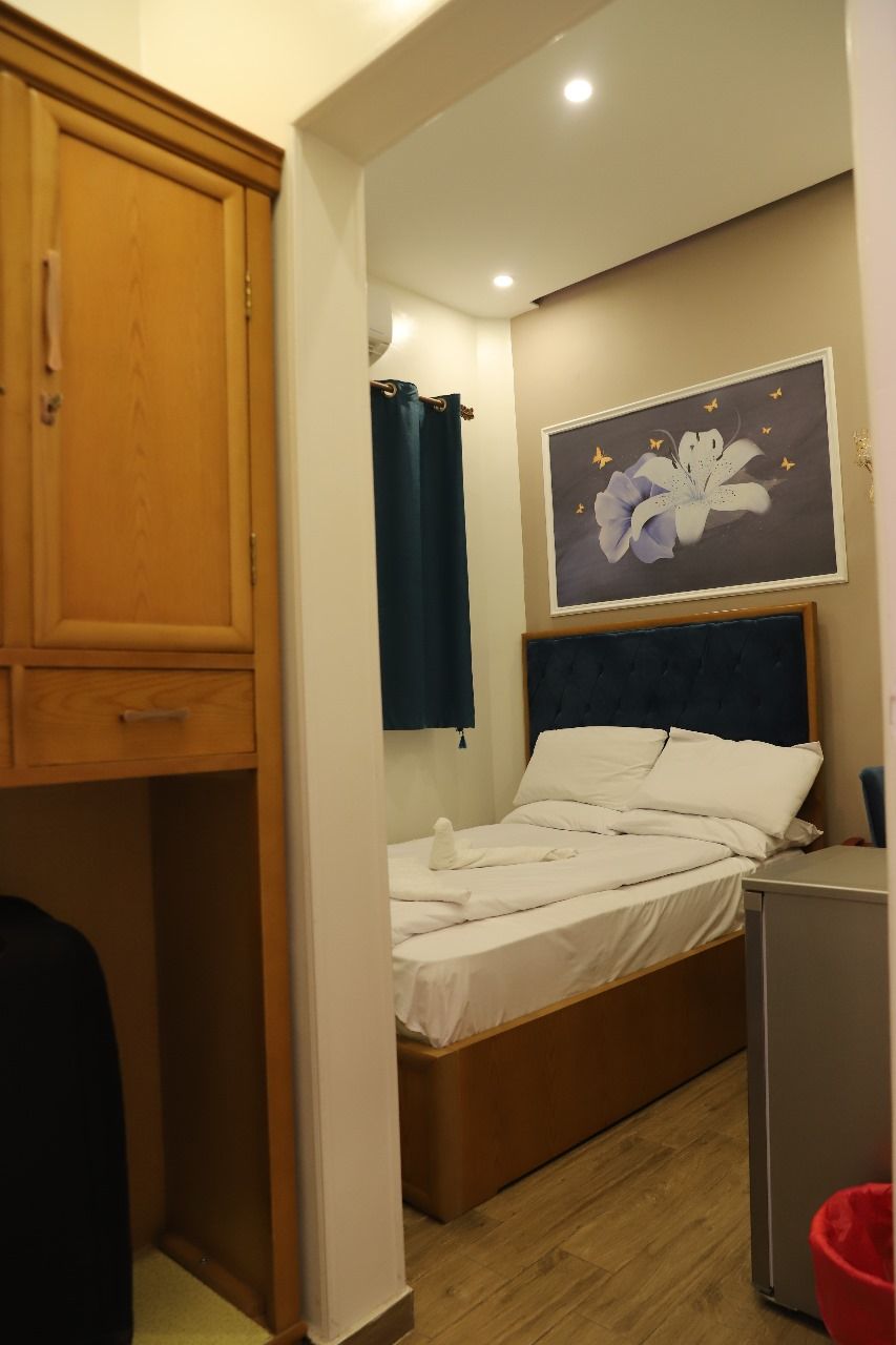 A bedroom with a bed, a wooden wardrobe, a small desk, a red trash bin, and a framed floral artwork on the wall.
