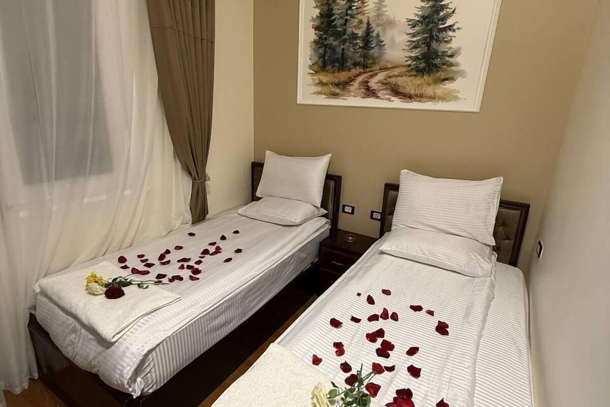 Two single beds with white linens, a painting of trees on the wall, and a curtain on the window.