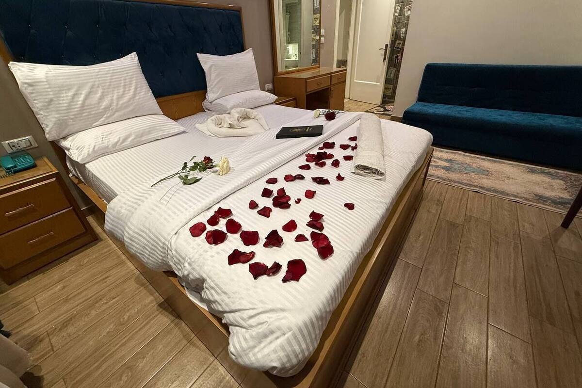A bed with white linens, a blue headboard, and red rose petals scattered on the bed.