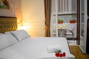 Une chambre d’hôtel avec un lit, une petite table et des chaises, un balcon agrémenté de plantes en pot et une vue sur la rue.