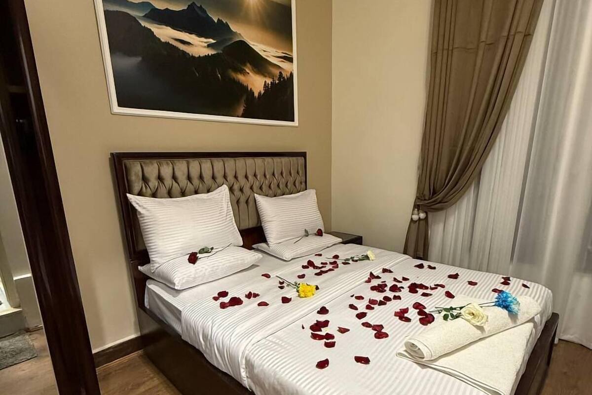 A bedroom with a large bed, a headboard, a painting of mountains, and a window with curtains.