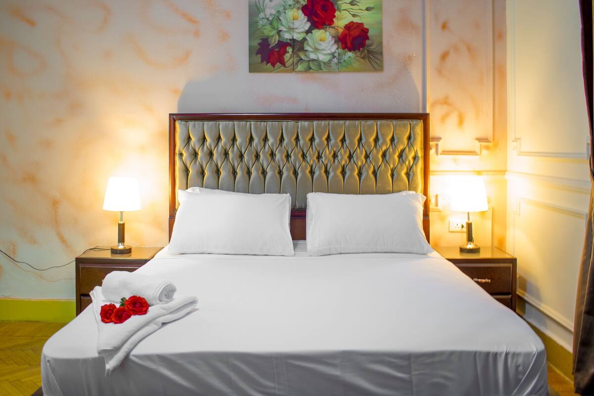 A neatly made bed with white linens, two bedside lamps, and a painting of flowers on the wall.