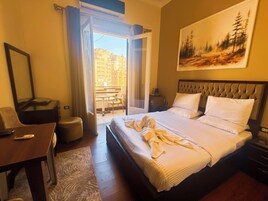 Habitación de hotel con cama, escritorio, silla, una mesita, ventana con cortinas y un cuadro en la pared.