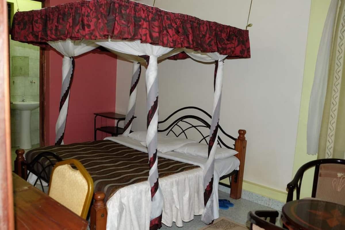 A single bed with a canopy, a wooden chair, a small table, and a sink in the background.