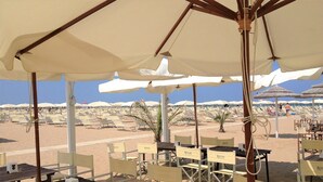 Un área de estar al aire libre con mesas y sillas bajo sombrillas, con vistas a una playa arenosa con sillones y sombrillas al fondo.