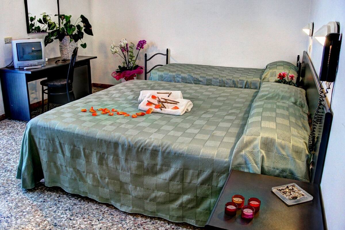 A hotel room with two beds, a desk with a computer, a chair, a small table with candles, and a plant.