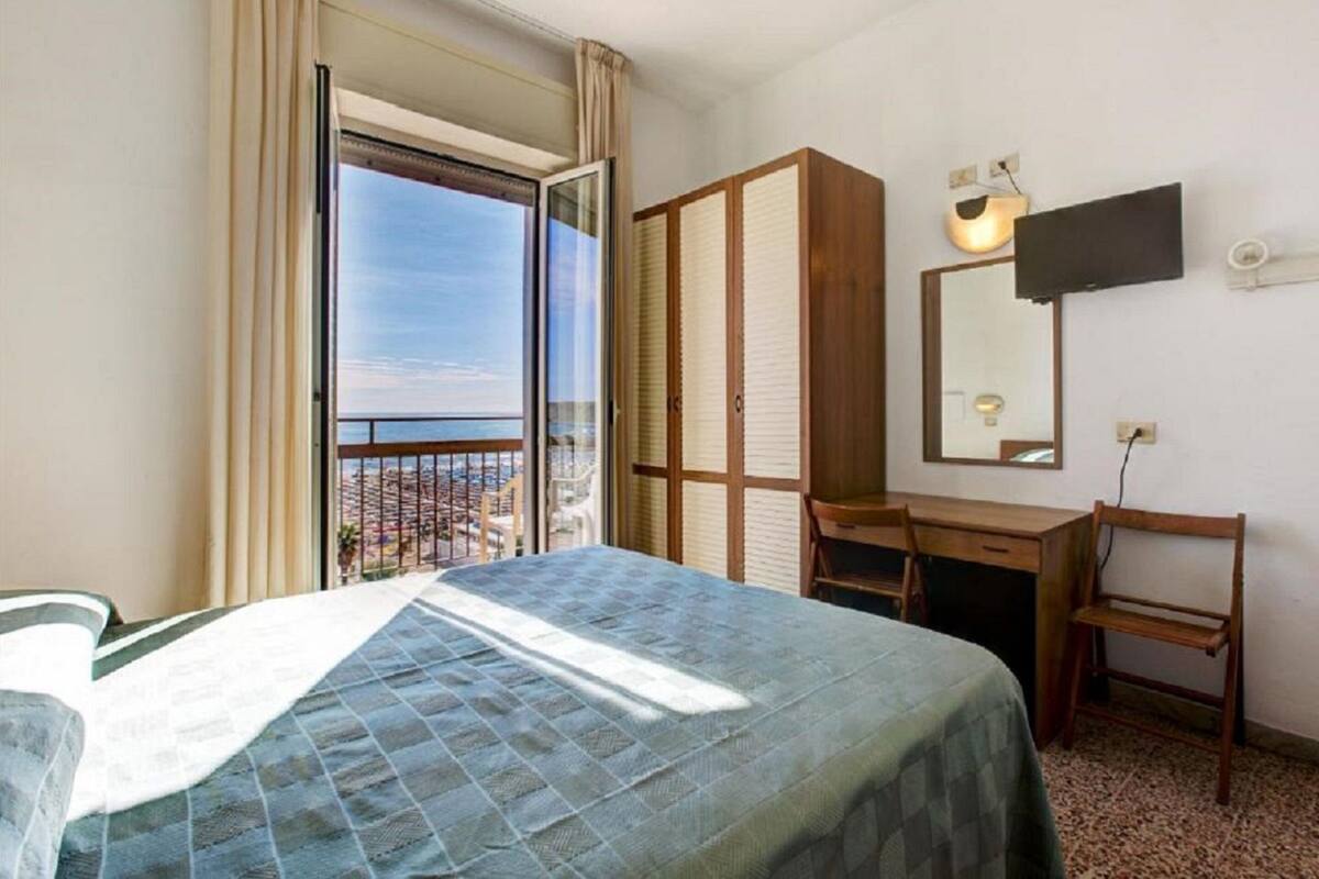 A hotel room with a bed, a desk, a chair, a wardrobe, a television, and a balcony with a view.