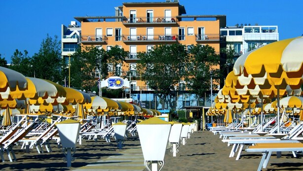 Una playa con filas de tumbonas y sombrillas, cielo despejado y un edificio con balcones al fondo.